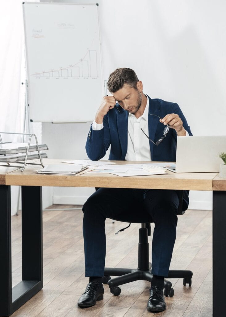 Male crisis manager in a suit sitting behind table, looking at papers scattered across his desk with one hand supporting his head at the temple