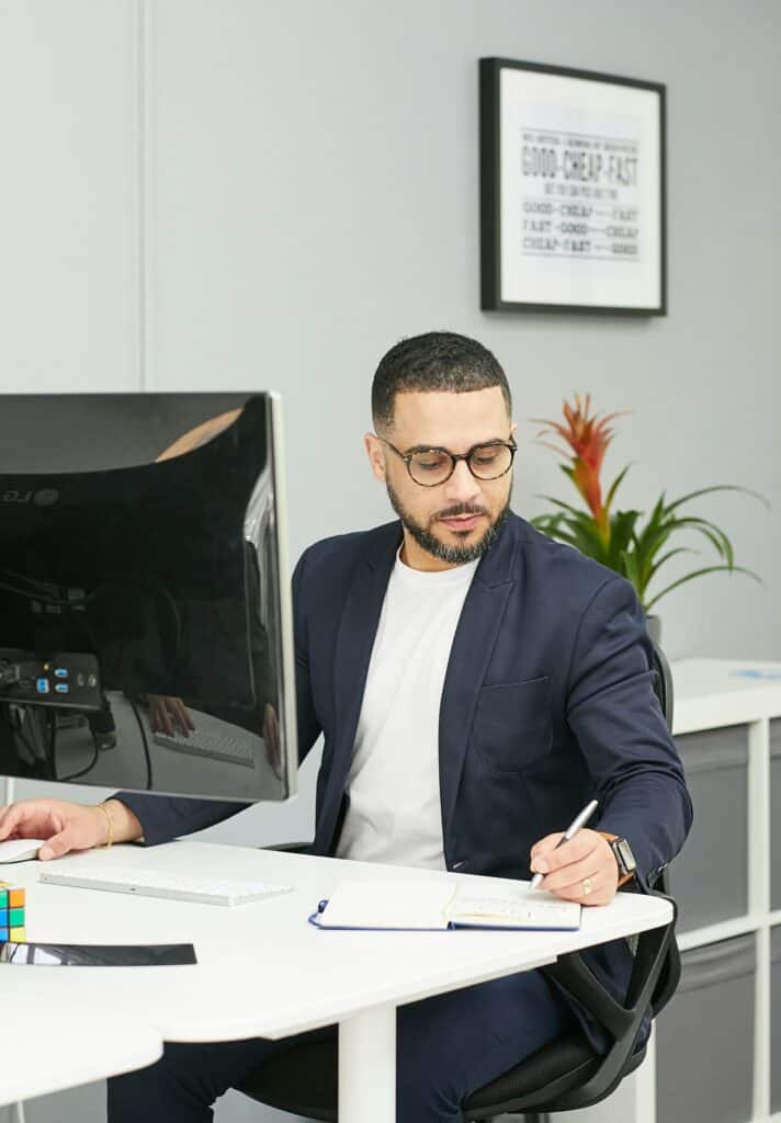 Operations manager in a suit sitting at is desk behind computer, taking notes in his notepad