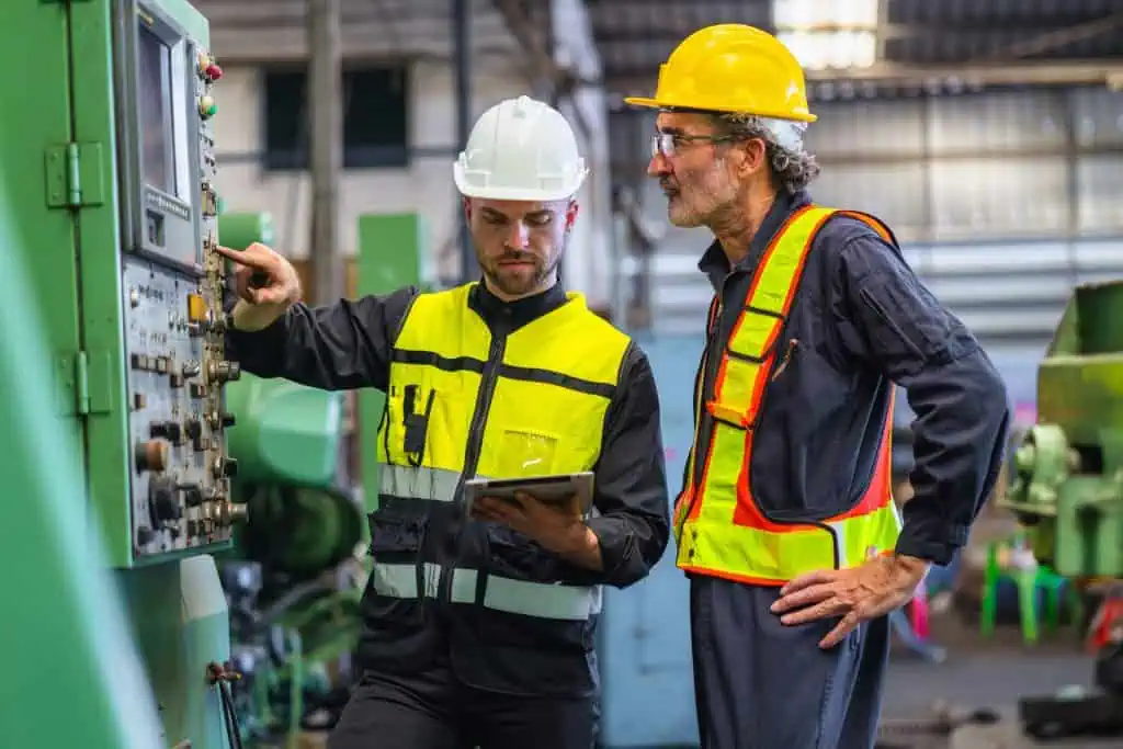 Two men in safety gear are standing next to a machine, one pointing at a button, they are discussing safety