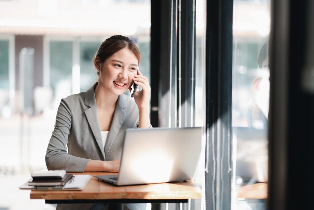 Young asian business woman talking on smartphone with laptop in front of her at a desk in a room with big windows.