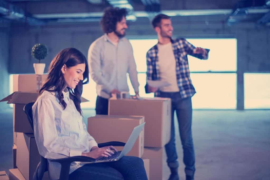 Three relocation management experts in empty building, two men stand in the backround behind boxes, woman is sitting on the ground with laptop in her lap
