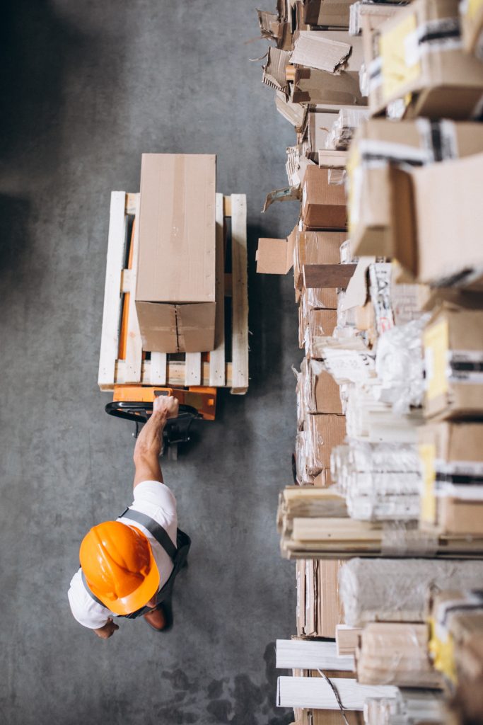 Warehouse worker with protective orange helmet dragging a loaded card behind him, shown from above view
