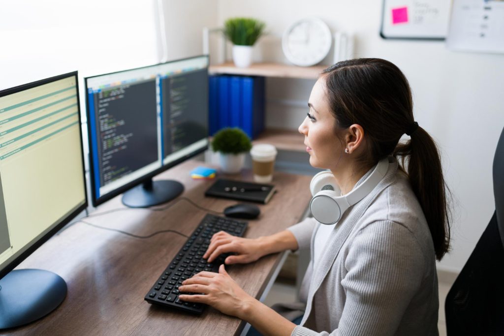 Young woman, IT integration services expert, typing on the computer and writing a code for a software app, using two monitors